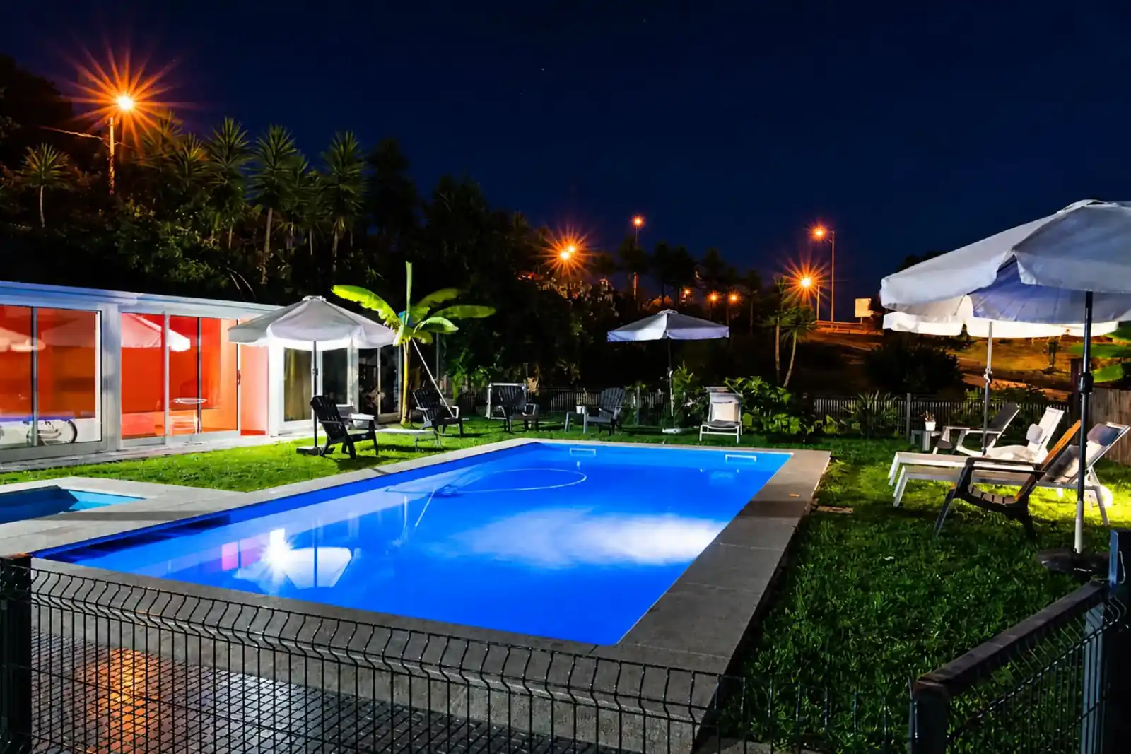 A piscina exterior iluminada à noite, com espreguiçadeiras e chapéus de sol