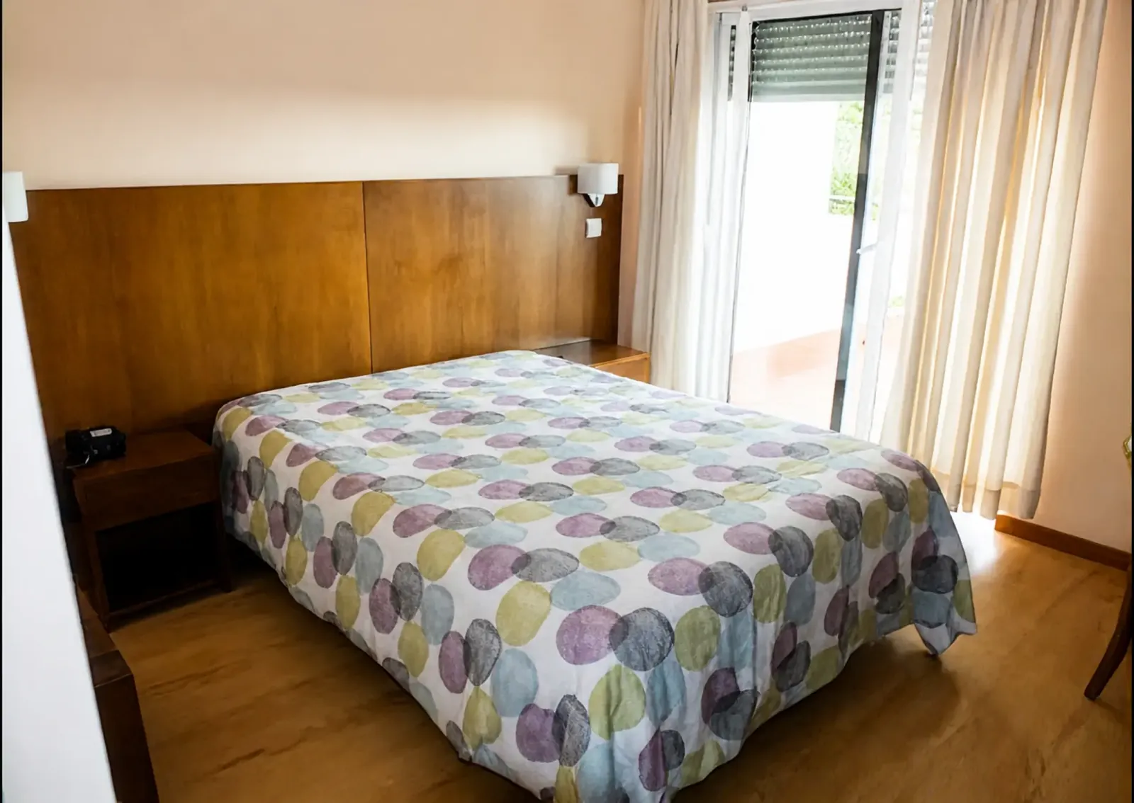 Quarto duplo com cabeceira de madeira e portas de varanda a deixar entrar a luz do dia
